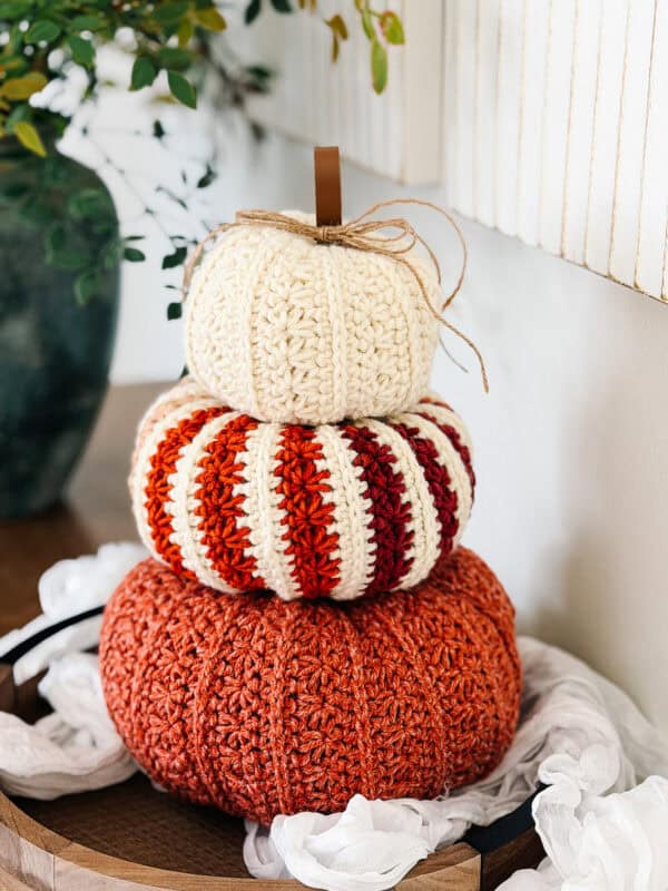 Three crocheted pumpkins in cream, striped orange and red, and solid orange are stacked on a wooden surface near a white cloth and green plant.