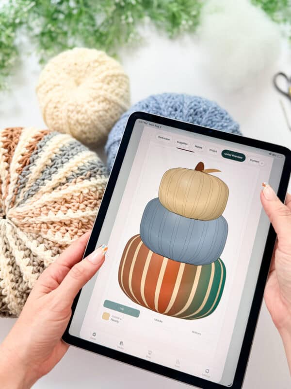 A person holds a tablet displaying a digital illustration of three stacked pumpkins; yarn pumpkins and yarn are on the table nearby.