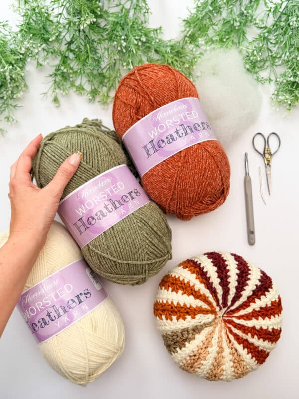 Three skeins of worsted yarn, a crochet hook, scissors, stuffing, and a striped crochet piece are arranged on a white surface with green foliage at the top.