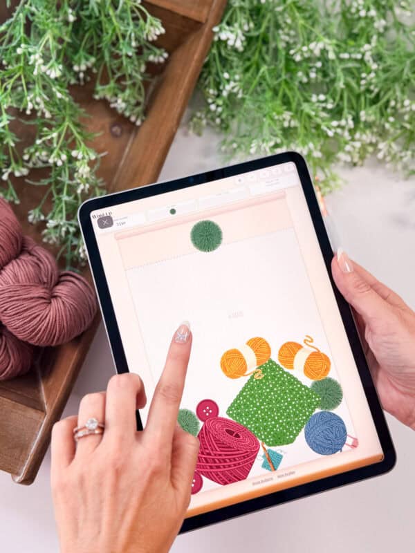 A person uses a tablet displaying a digital design with yarn and fabric graphics, possibly interacting with a crochet game, surrounded by yarn balls and leafy green artificial plants.