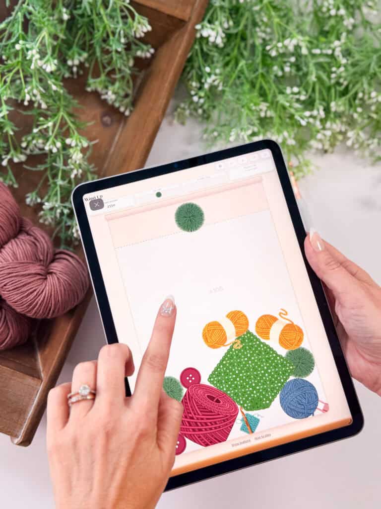 A person uses a tablet displaying a digital design with yarn and fabric graphics, possibly interacting with a crochet game, surrounded by yarn balls and leafy green artificial plants.