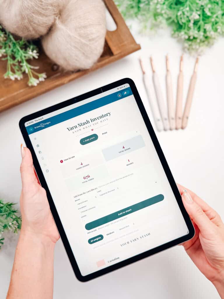 Person holding a tablet displaying a “Yarn Stash Inventory” app, with yarn balls, greenery, and crochet hooks visible in the background on a white surface—perfect for crafters looking to organize their supplies or use a handy crochet yarn calculator.