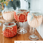Four wine glasses with knitted covers in cream and rust colors, displayed on a wooden table. A label advertises a free pattern and video tutorial from brianakdesigns.com.