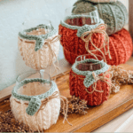 Four wine glasses with knitted, pumpkin-themed covers in cream and orange, placed on a wooden surface with pinecones and stacked knitted pumpkins in the background.