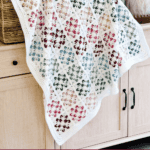 A multicolored blossom granny square crochet blanket draped over a light wooden cabinet, with a basket and vase in the background.