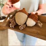 Person holding a tray with three crocheted acorns in different sizes and colors, decorated with small branches. Text at bottom reads, “Wheat Stitch Acorns - 3 Sizes.”.