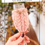 A hand holds a wine glass adorned with a chunky pink wheat stitch knitted cover, reminiscent of a crochet wine glass cover, and tied with a twine bow. The background is softly blurred with green and yellow foliage.