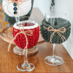 Three wine glasses with knitted covers in white, red, and green, each tied with a bow, displayed on a wooden surface. Text reads: “Wheat Stitch Wine Glass Cover.”.