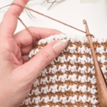 A person’s hand holds a wooden crochet hook, working on a two-toned tan and white project featuring a houndstooth stitch pattern that would beautifully complement any crochet acorn pattern. The image includes text reading “How to Crochet the Houndstooth Stitch.”.
