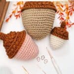 Three crocheted acorns in various sizes are displayed with a crochet hook, scissors, stitch markers, and autumn leaves on a white surface.