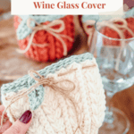 Hand holding a crocheted wheat stitch wine glass cover tied with twine, with other covers and an empty wine glass on a wooden table.