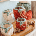 Four wine glasses with knitted, pumpkin-inspired covers sit on a wooden tray, surrounded by dried botanicals and crocheted pumpkin decorations in the background.