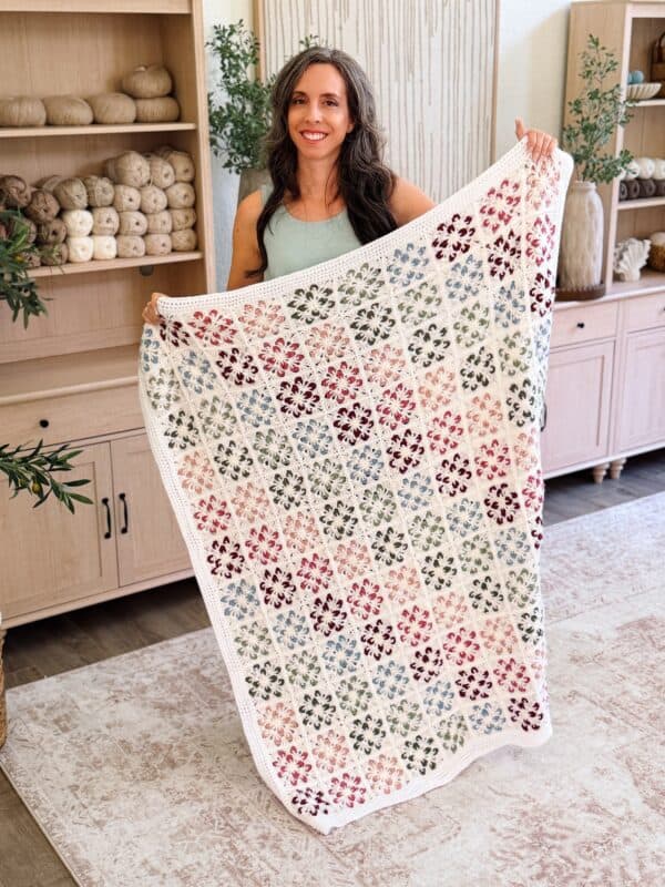 Woman holding a crocheted blanket with a colorful floral pattern, standing in a room with wooden shelves and yarn in the background.
