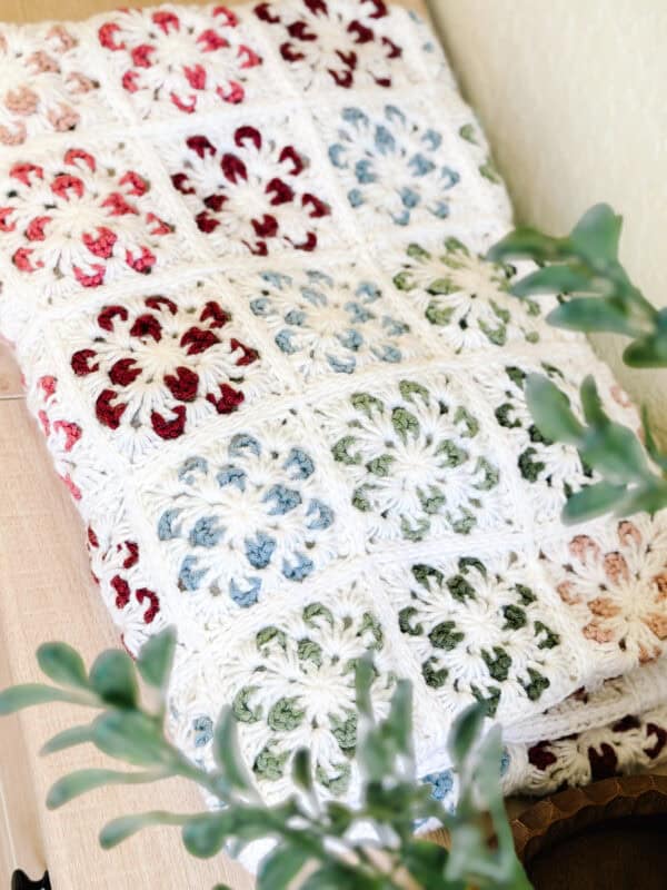 A crocheted blanket with multicolored granny squares, mostly white with red, blue, green, and beige accents, is folded on a wooden surface next to green leafy plants.