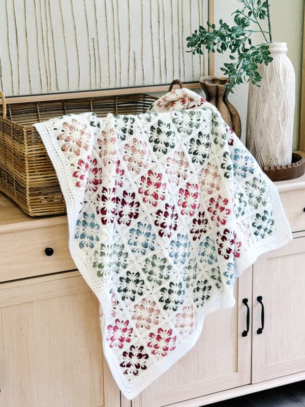 A multicolored crocheted blanket with a floral pattern is draped over a light wood cabinet next to a basket, vase, and decorative branch.