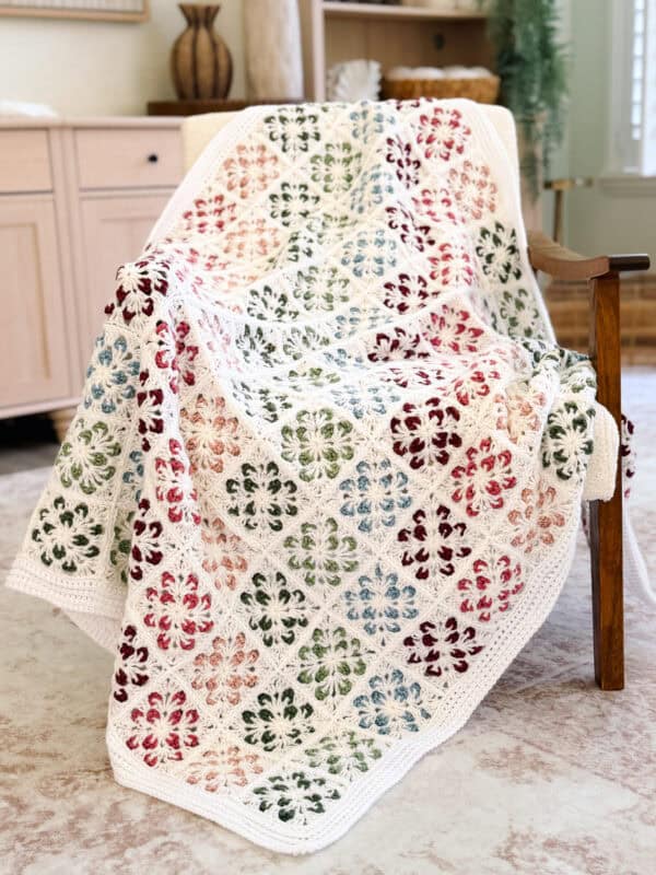 A crocheted blanket with a white base and multicolored floral square motifs draped over a wooden chair in a cozy room.