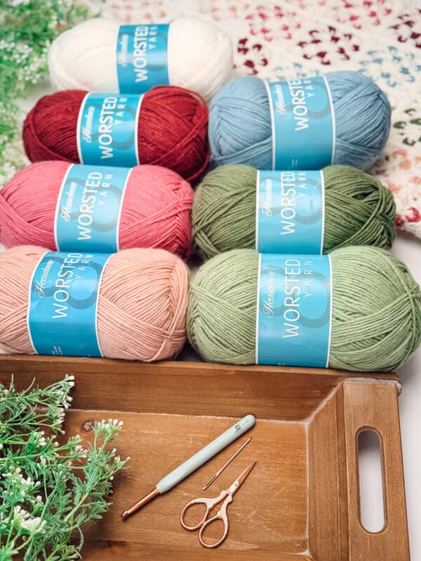Six skeins of worsted yarn in various colors, a crochet hook, and scissors are arranged on a wooden tray with green foliage and a crocheted blanket in the background.
