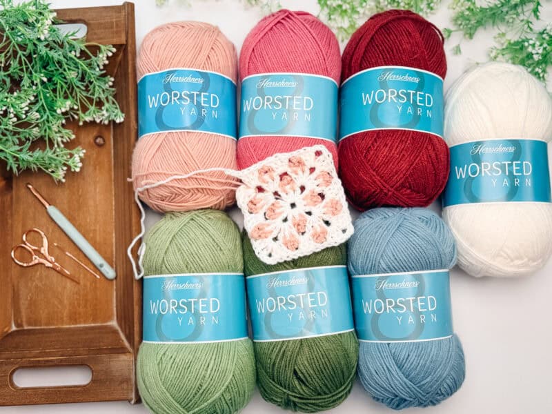 Eight balls of worsted yarn, a crochet hook, scissors, and a crochet square are arranged on a tray and table with green plants.