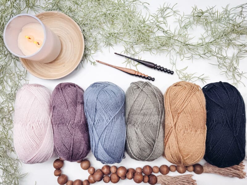 Six skeins of yarn in blush, plum, denim blue, taupe, camel and black lined up on a white surface with two crochet hooks, a candle, wooden beads and greenery, ready for practicing the houndstooth stitch