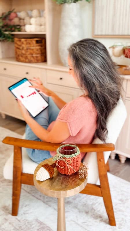 A woman with long gray hair sits in a chair using a tablet, perhaps enjoying a crochet solitaire game. In the foreground, a small table holds yarn pumpkins and knitted decor.