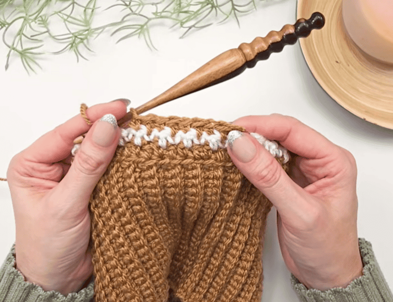 Two hands holding a brown crocheted item with a row of white stitches, using a wooden crochet hook—perhaps working on a charming crochet acorn pattern. A plant and a candle are visible in the background.