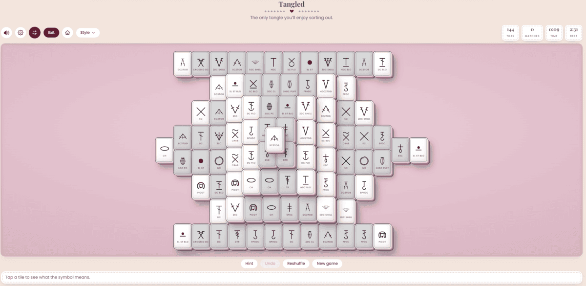 A digital crochet solitaire game board with tiles displaying unfamiliar symbols is shown on a pink background. Game controls and menu options are visible at the top and bottom.