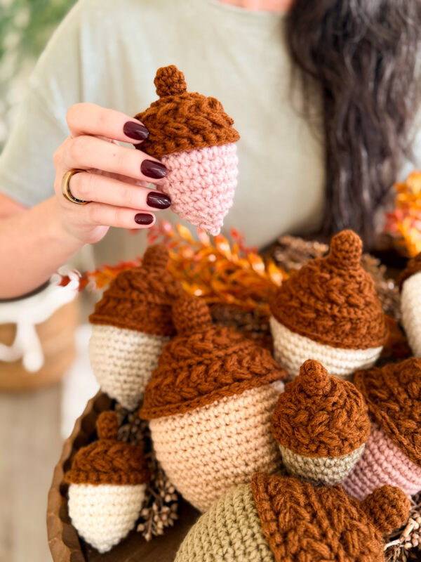 A person with dark nail polish holds a crocheted acorn above a basket filled with similar handmade, brown-topped acorns.