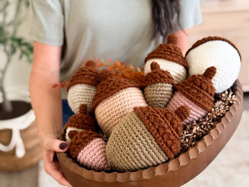 Person holding a wooden bowl filled with large, crocheted acorns in various shades of brown, beige, and pink.