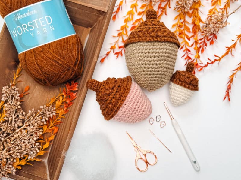 Three crocheted acorns in different sizes with brown tops sit near fall leaves, a ball of brown yarn, crochet hook, scissors, needle, stitch markers, and fiberfill stuffing on a white surface.