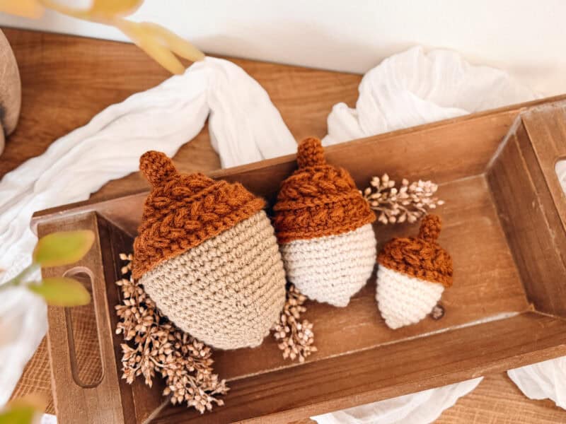 Three crocheted acorns with brown tops and beige bottoms are placed on a wooden tray with dried flowers, atop a wooden surface.