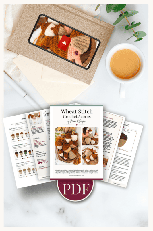 Flat lay of a crochet acorn PDF pattern, printed pages, a coffee cup, envelope, greenery, and a tablet showing a video tutorial thumbnail.