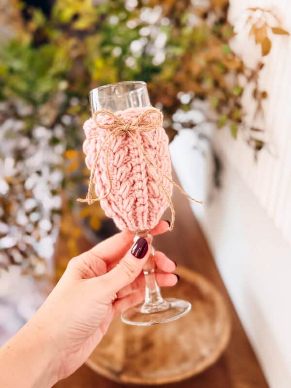 A hand holds a champagne glass wrapped in a crochet wine glass cover, knitted in pink and tied with twine. Green leafy plants are blurred in the background.
