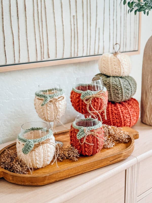 Three wine glasses wrapped in knitted covers and three stacked knitted pumpkins rest on a wooden tray with pinecones, sitting on a light wood surface below framed artwork.