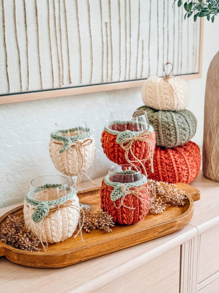 Three wine glasses wrapped in knitted covers and three stacked knitted pumpkins rest on a wooden tray with pinecones, sitting on a light wood surface below framed artwork.