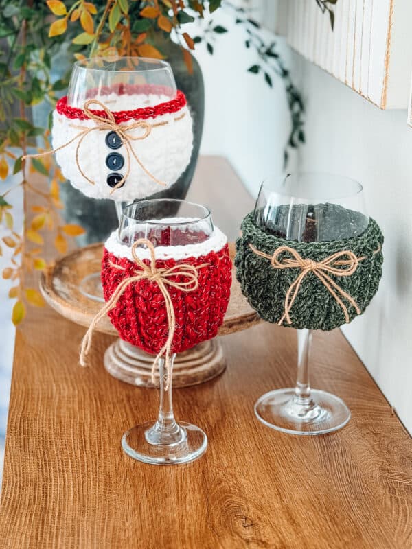 Three wine glasses with knitted cozies in red, white, and green, each decorated with twine bows and buttons, displayed on a wooden surface.