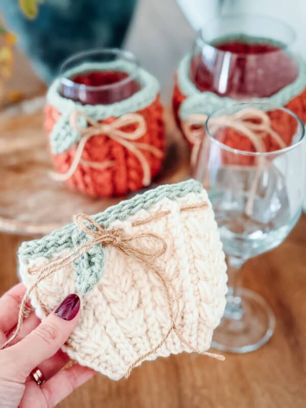 Hand holding a cream-colored knitted cover with a bow, next to wine glasses and glasses wrapped in orange and green knitted covers.
