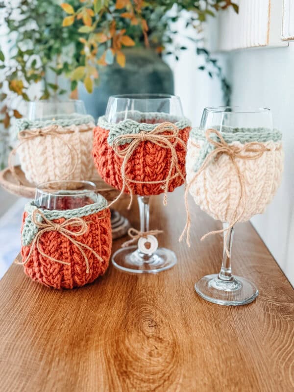 Four wine glasses on a wooden surface, each covered with a knitted cozy in cream or orange, tied with twine bows. Greenery is blurred in the background.