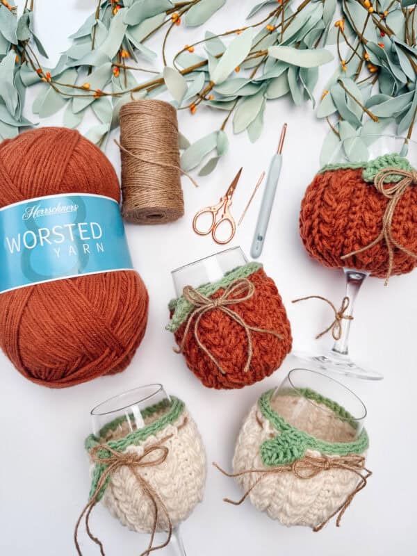Four wine glasses with knitted cozy covers, a skein of rust yarn, twine, scissors, a crochet hook, and faux eucalyptus branches on a white surface.