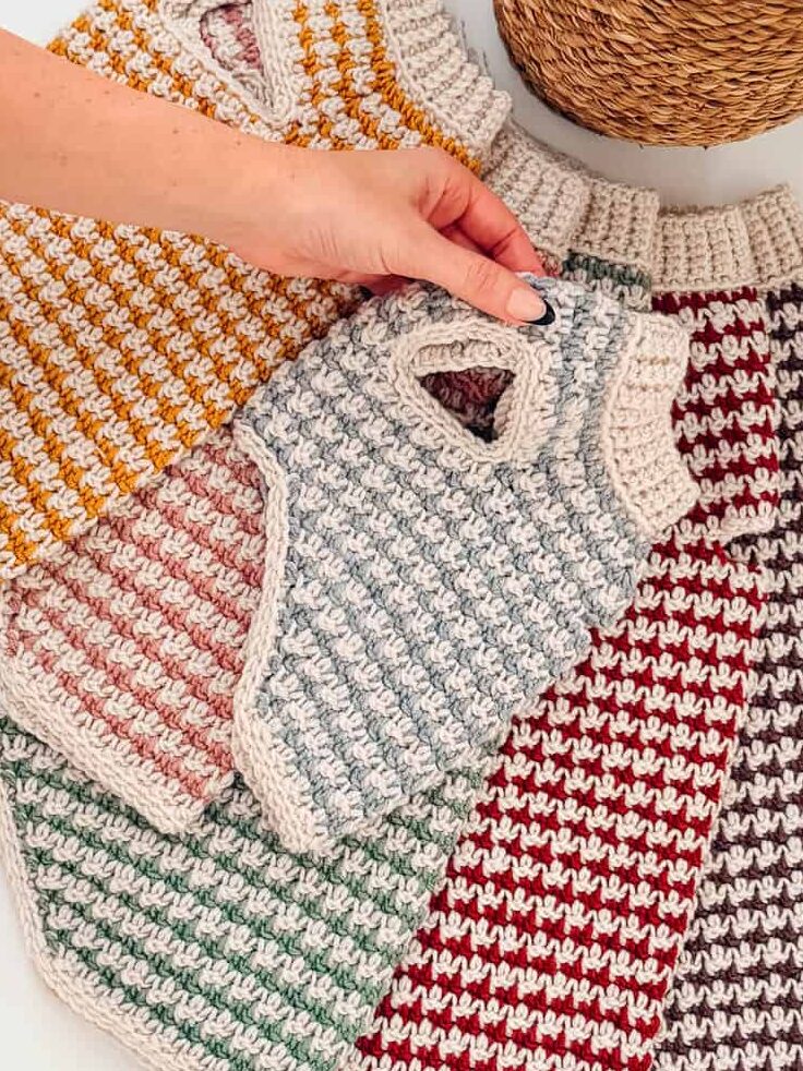 A hand arranging a stack of five small, crochet dog sweaters in various striped colors.