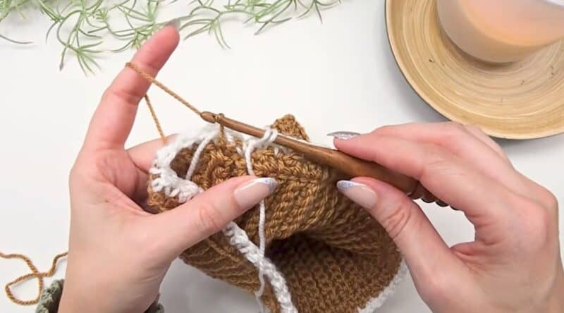 Hands working camel yarn on top of a cream round of the houndstooth stitch in the round, with the cream strand running up the inside of the work