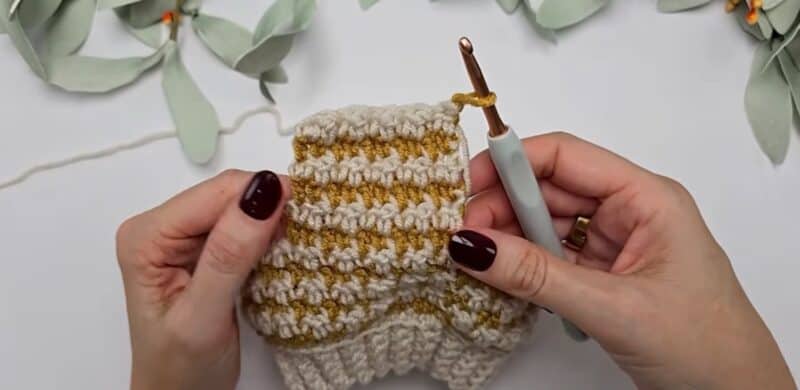 A flat cream and mustard houndstooth crochet panel with the mustard yarn on the hook at one edge and the dropped cream yarn trailing off the opposite edge