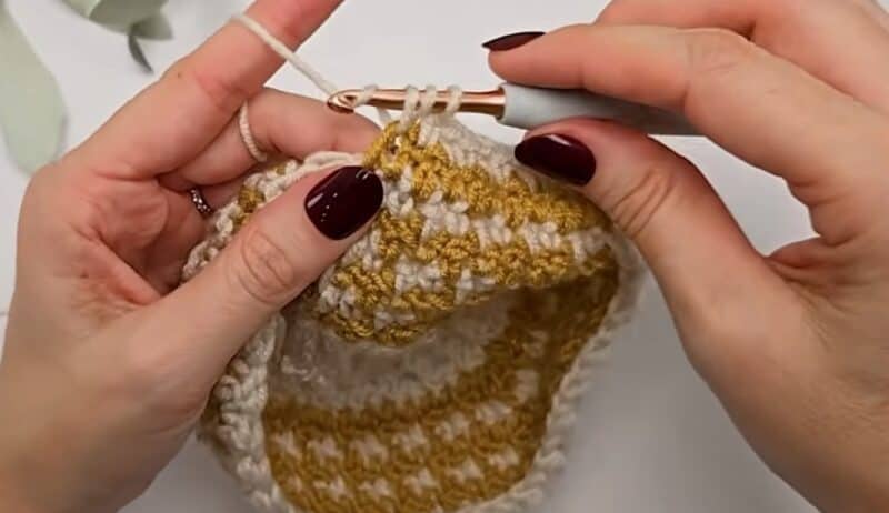 Hands working a double crochet in cream yarn around a mustard and cream houndstooth crochet tube, with several loops on the hook