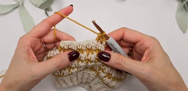 Hands starting a new row of the houndstooth stitch in mustard yarn on top of a cream row, with the hook at the edge of the flat panel