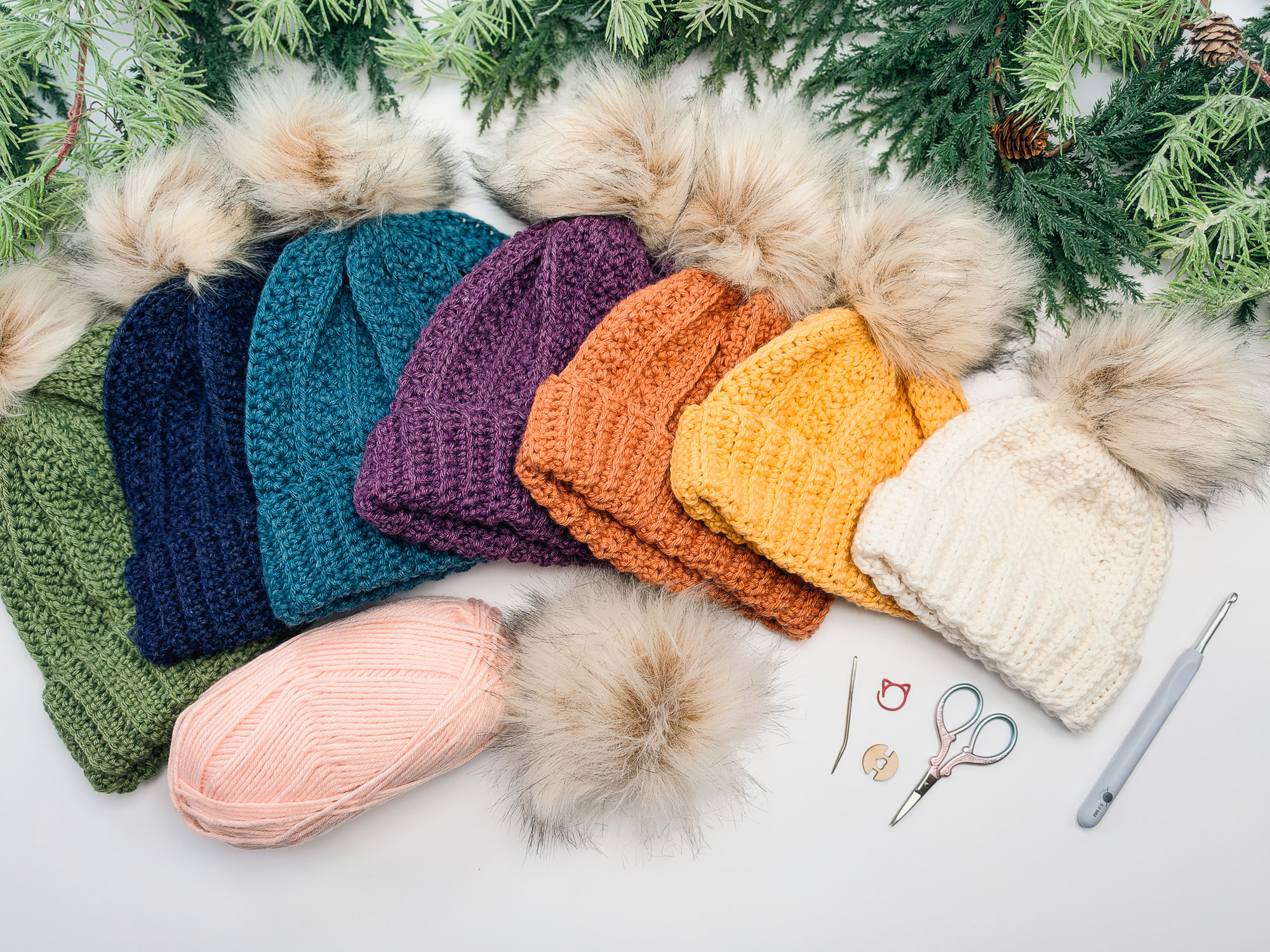 Petal Loop Crochet Beanies in green, navy, teal, purple, orange, mustard yellow and cream with faux fur pom-poms, laid out with pink yarn, a crochet hook, scissors, stitch markers and a yarn needle