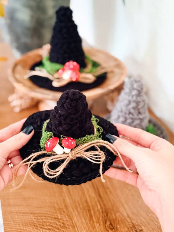 Hands hold a mini crochet witch hat decorated with green moss, a jute twine bow, and two small red-and-white mushrooms.