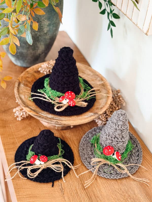 Three mini crochet witch hats in black and grey, decorated with green moss, twine bows, and miniature red toadstools.