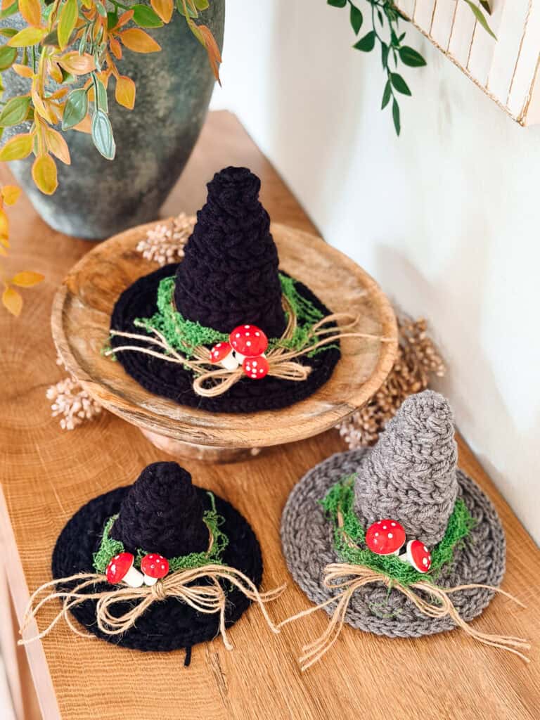 Three mini crochet witch hats in black and grey, decorated with green moss, twine bows, and miniature red toadstools.