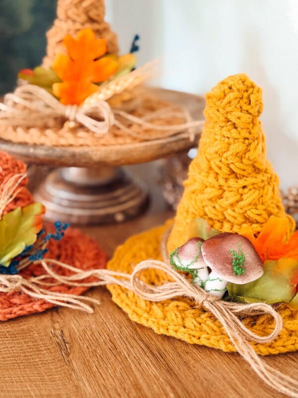 Three mini crochet witch hats in autumn colors, decorated with twine bows, artificial leaves, and miniature mushrooms.