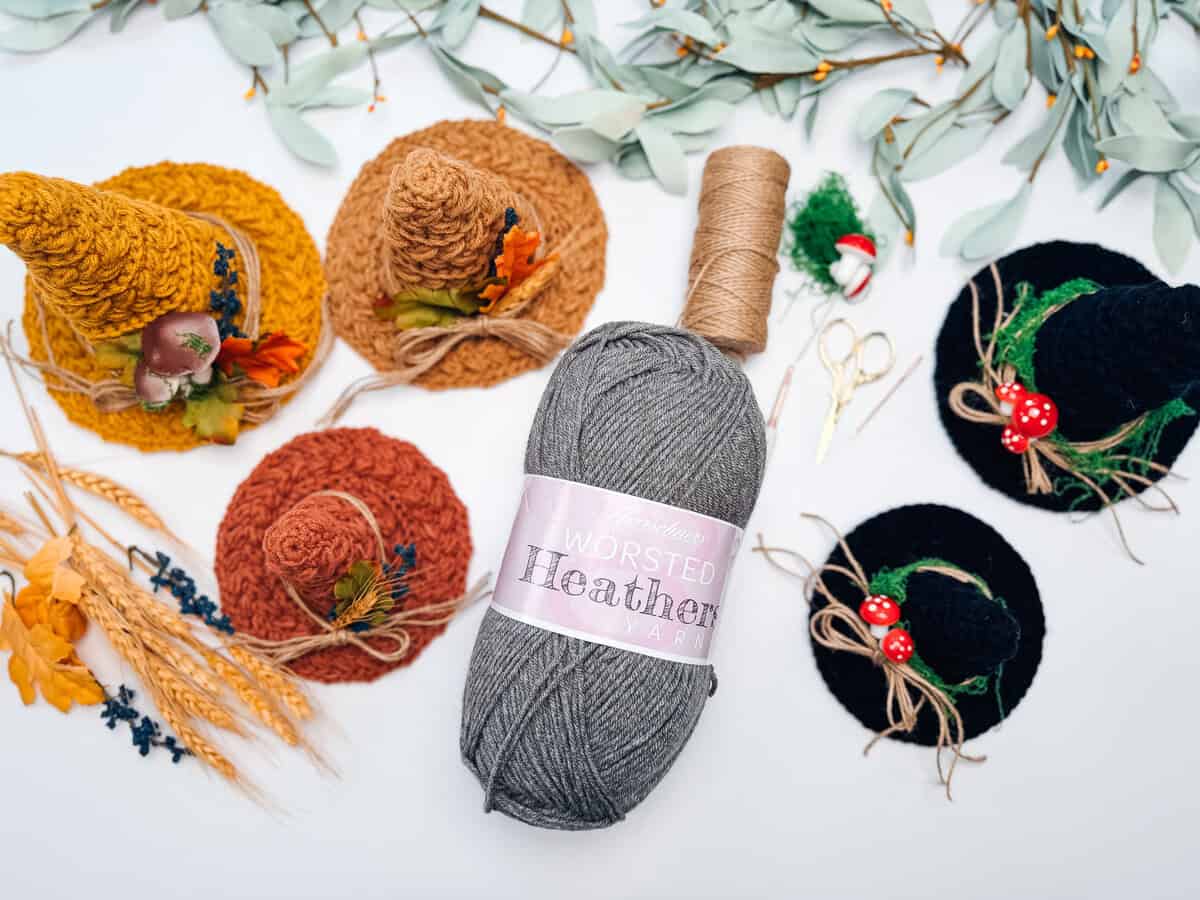 A skein of grey worsted yarn surrounded by mini crochet witch hats in orange, yellow, and black, alongside decorative wheat, twine, and miniature mushrooms.