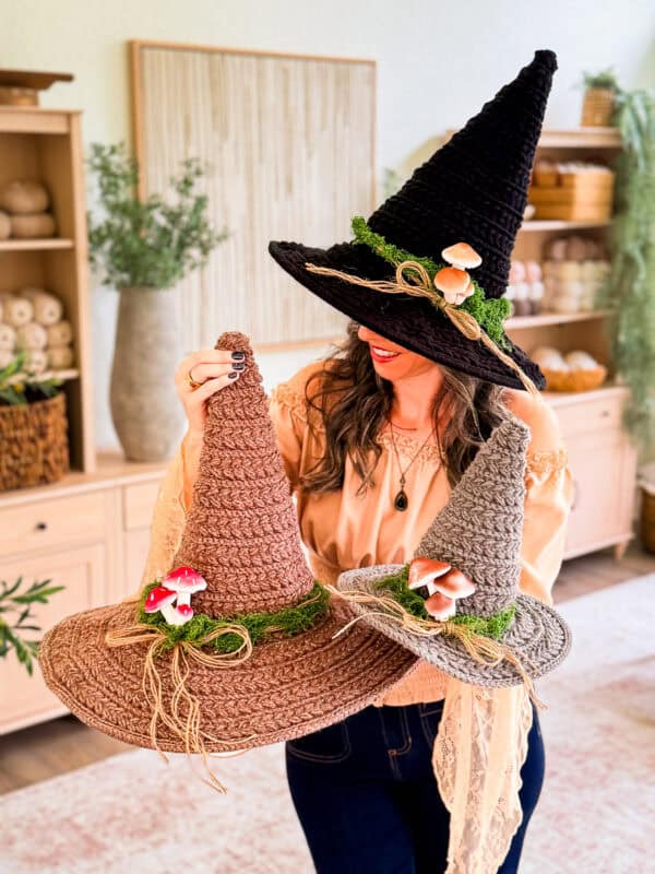 A person holding three crocheted witch hats decorated with faux moss, twine, and mushrooms, crafted using a crochet witch hat pattern.
